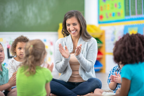 A female teacher is sitting on the floor with her group of students at a classroom. They are all singing and clapping their hands together to a song.